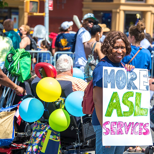 A parade-goer at the Disability Parade 2024 holds a sign reading: “More ASL Services.”